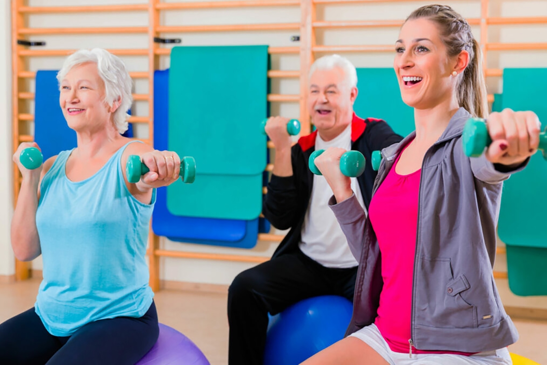 Sportgruppe: Trainerin und zwei Kursteilnehmer mit je zwei kleinen Hanteln sitzen auf Fitnessbällen